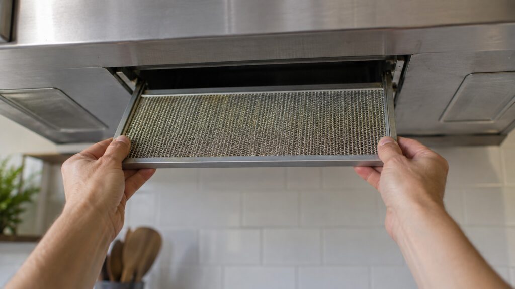 A metal mesh grease filter being slid out of the underside of an over-the-range microwave, with the vent opening visible above it